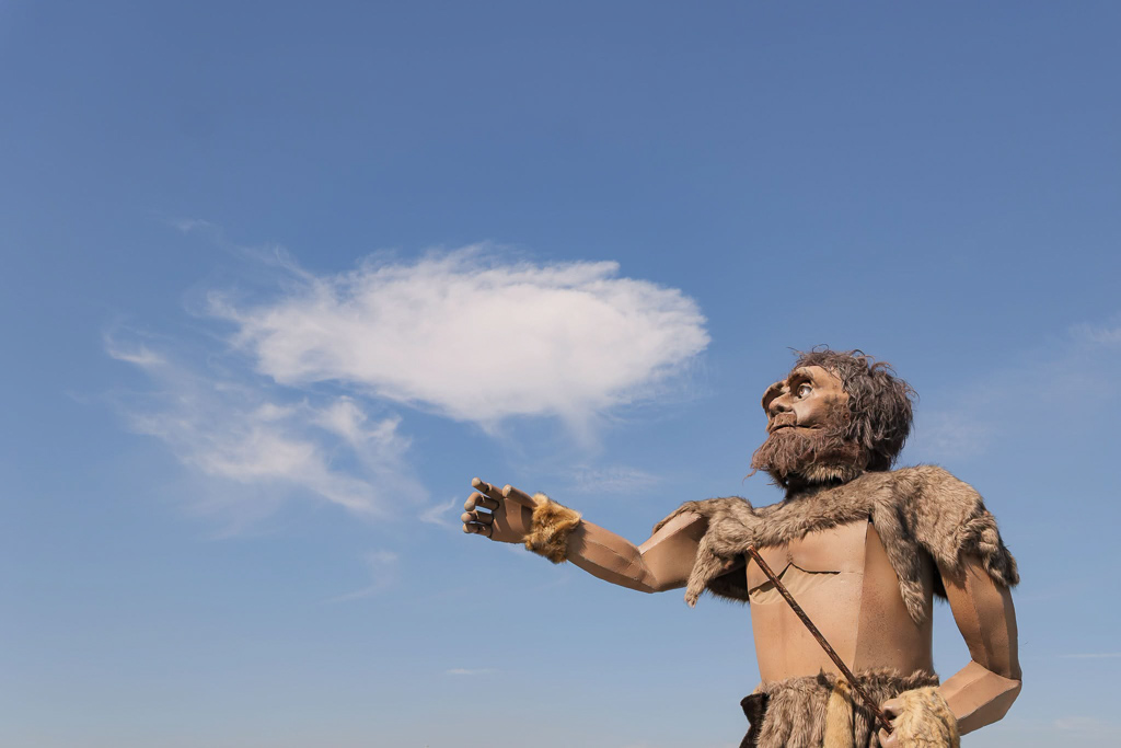 Personagem pré-histórica de braço estendido, contra o céu azul.